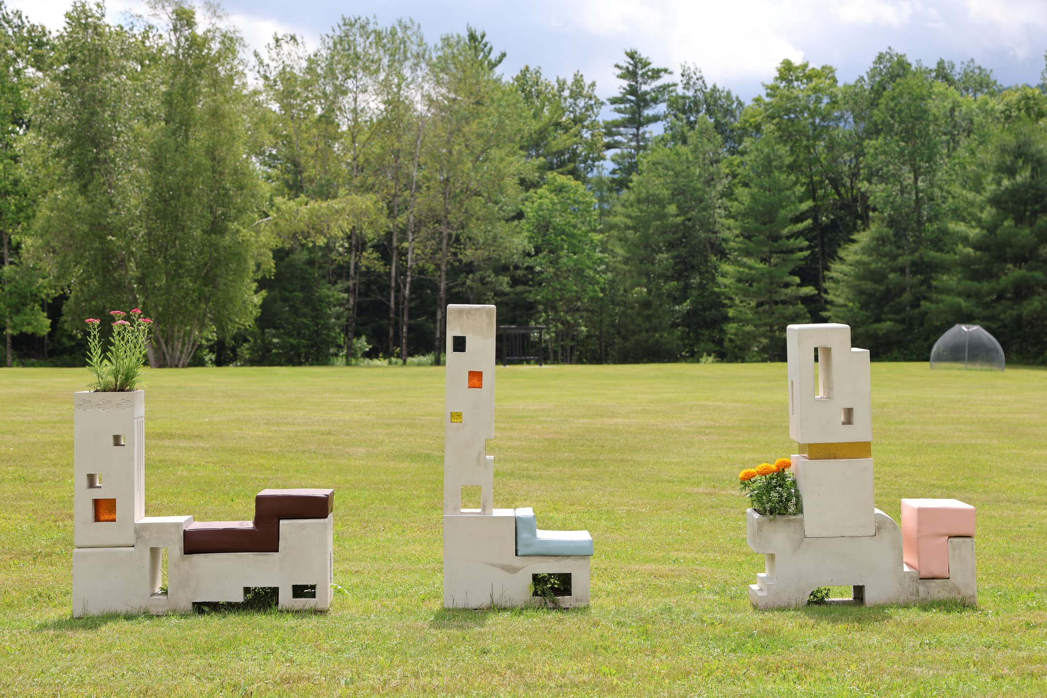 Three concrete sculptures made to look like chairs are situated in a line in a large field.