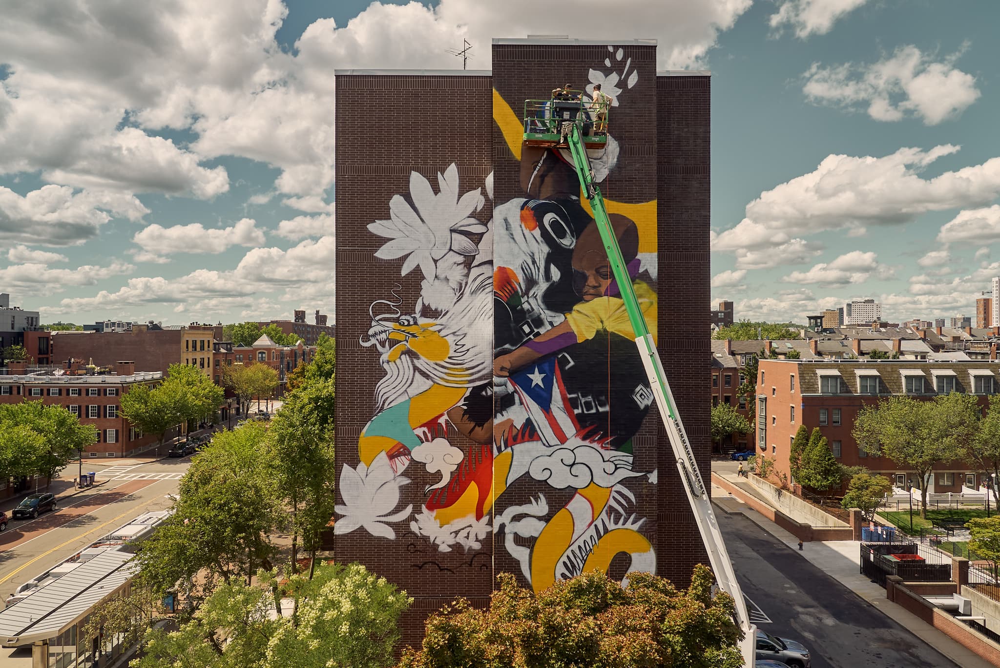 Victor "Marka27" Quiñonez's mural in the city, set against the backdrop of a blue sky filled with clouds.