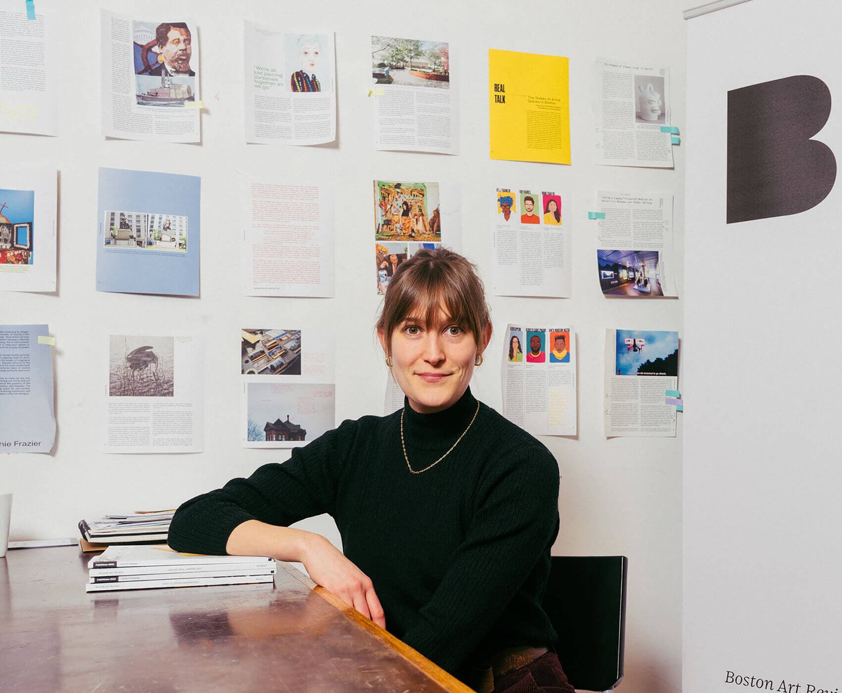 Jameson Johnson, a light-skinned woman with bangs and hair tied into a pony tail, sits at a table with her elbow rested atop a stack of magazines, smiling at the camera.