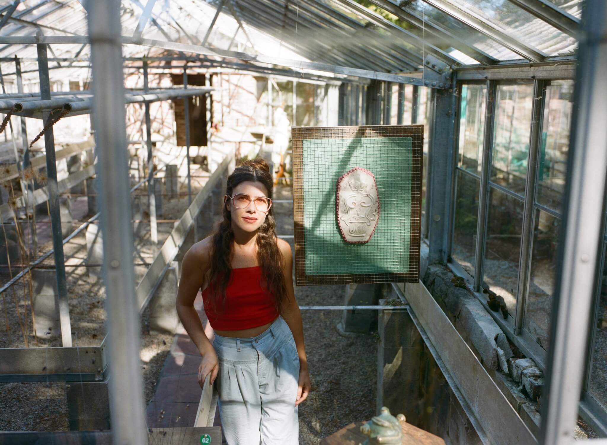 An artist stands beside a hanging piece of art in a greenhouse.